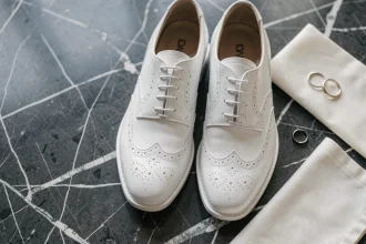 Chaussures derby blanches en cuir pour mariage posées sur marbre noir avec alliance et pochette ivoire